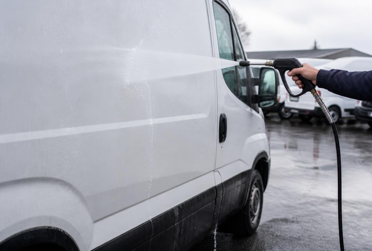 Pressure washer trigger gun rinsing the side of a white commercial work van in a fleet parking lot