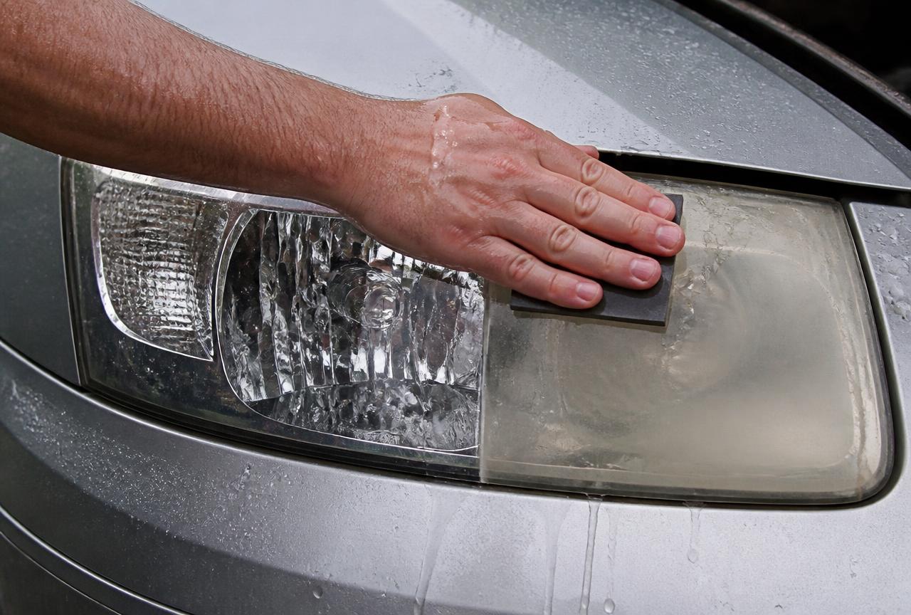Wet sanding block clearing a yellowed and hazy car headlight lens, half restored to clear